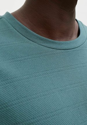 Close-up van een donkerhuidig persoon met een teal textuurstof T-shirt met ronde hals, met focus op de halslijn en het bovenste borstgedeelte.