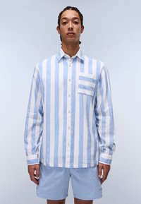 Camisa de botones de manga larga a rayas azul claro y blanca con un bolsillo en el pecho, hecha de una tela texturizada, combinada con pantalones cortos azul claro.