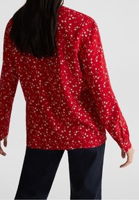 Femme portant un chemisier rouge à manches longues avec un petit imprimé floral blanc, associé à un pantalon noir, vue de dos.