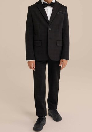 Dreng iført sort jakkesæt med butterfly, hvid skjorte og sorte sneakers, stående mod en ensartet beige baggrund.