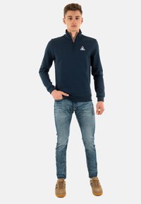 Navy pullover with a half-zip collar, ribbed cuffs, and a logo. Paired with faded blue jeans and brown sneakers with black accents.