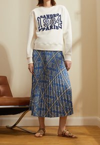 Kermainen collegepaita, jossa sininen "SANDRO 1984 PARIS" -kuvio. Virtaava sininen hame, jossa valkoinen paisley-kuvio. Ruskeat sandaalit, joissa on pyöreitä yksityiskohtia.