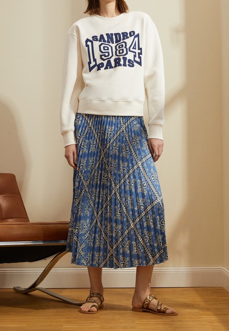 Kermainen collegepaita, jossa sininen "SANDRO 1984 PARIS" -kuvio. Virtaava sininen hame, jossa valkoinen paisley-kuvio. Ruskeat sandaalit, joissa on pyöreitä yksityiskohtia.