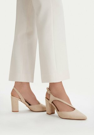 Kobieta nosząca beżowe zamszowe sandały na słupku typu slingback, zestawione z białymi krótkimi spodniami, na białym tle.