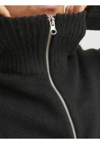 Zbliżenie dłoni poprawiających czarny prążkowany sweter z srebrnym zamkiem błyskawicznym przy wysokim kołnierzu.