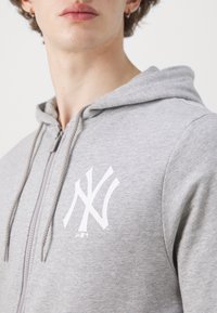 Haut de sweat à capuche zippé gris en tissu doux, présentant un grand logo blanc des New York Yankees sur la poitrine et une capuche avec cordon de serrage.
