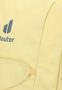 Gelber Stoff-Rucksack mit einer strukturierten Oberfläche, seitlichem Schlaufen und blauem Logo. Einfaches Design mit klaren Linien und funktionalen Akzenten.