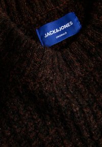 Suéter de punto marrón oscuro con un tejido texturizado y grueso. Presenta una etiqueta de tela azul con texto blanco, parcialmente visible en el área del cuello.