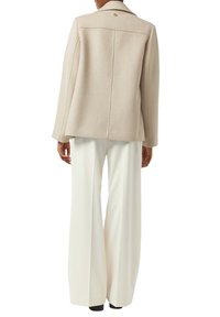 Beige, textured jacket with a tailored fit, featuring a back seam detail and a small logo above the collar. Paired with white, wide-leg pants.