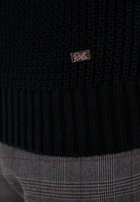 Czarny sweter dziany z prążkowanym dołem i małą brązową skórzaną metką z logo. Noszony na szarych spodniach w kratę z czerwonymi akcentami.