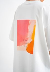 Witte oversized T-shirt met een grote grafische afbeelding op de achterkant, bestaande uit abstracte penseelstreken in roze, oranje en geel, samen met tekst.