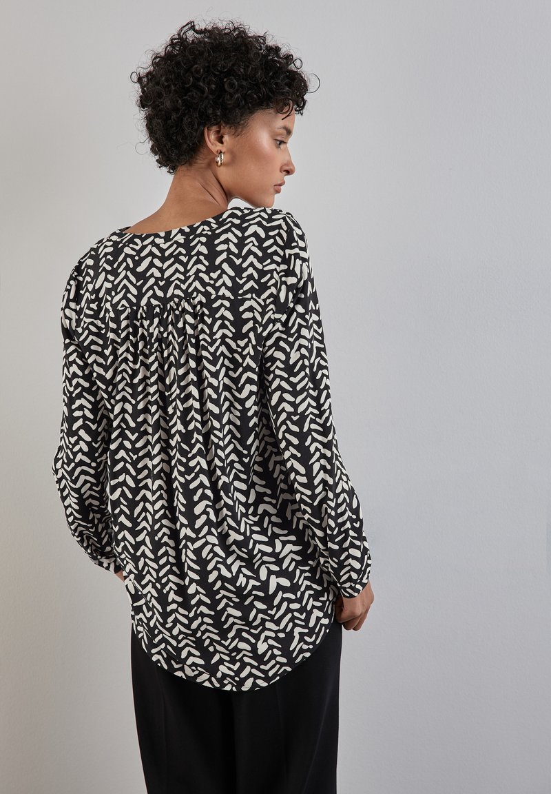 Blusa nera con motivo astratto bianco, tessuto leggero, vestibilità ampia, scollatura rotonda, dettaglio arricciato sul retro, maniche lunghe.