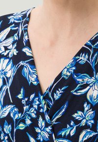 Blusa floral con escote en V profundo, con un fondo azul marino y vibrantes patrones botánicos en blanco y azul. Textura de tela suave evidente.