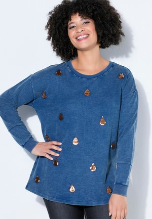 Uśmiechnięta kobieta o kręconych włosach, ubrana w niebieską koszulę z długim rękawem ozdobioną rozsypanymi miedzianymi cekinami.