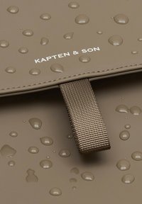 Waterdichte bruine tas met reliëflogo, met een gestructureerd oppervlak met zichtbare waterdruppels en een stoffen schouderband.