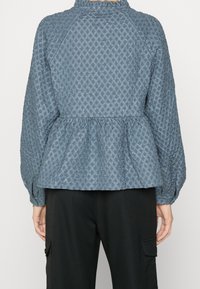 Giacca peplum blu con texture, orlo arricciato, colletto alto e maniche raglan, caratterizzata da un motivo a diamante e polsini elasticizzati.