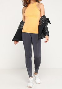 Débardeur de sport orange avec un col halter, associé à un legging gris foncé avec un design en chevrons, et une veste noire matelassée.