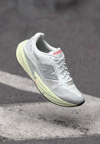 Der New Balance Laufschuh hat ein graues Mesh-Obermaterial mit reflektierenden Akzenten, eine gepolsterte gelbe Sohle und ein schlankes, leichtes Design.
