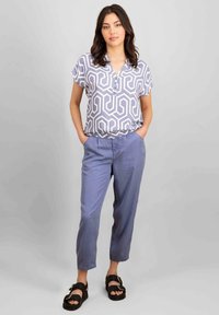Blusa leggera e stampata con design geometrico, maniche corte e una lista di bottoni, abbinata a pantaloni blu e sandali neri.