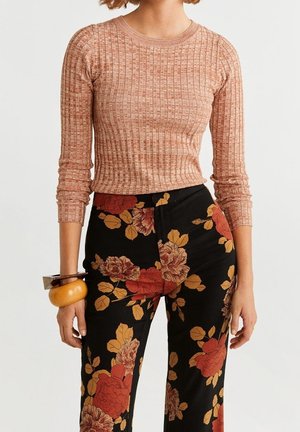 Kvinde iført en ribbet, lysebrun langærmet top og sorte bukser med store røde og gule blomsterprint, accessorized med chunky armbånd.