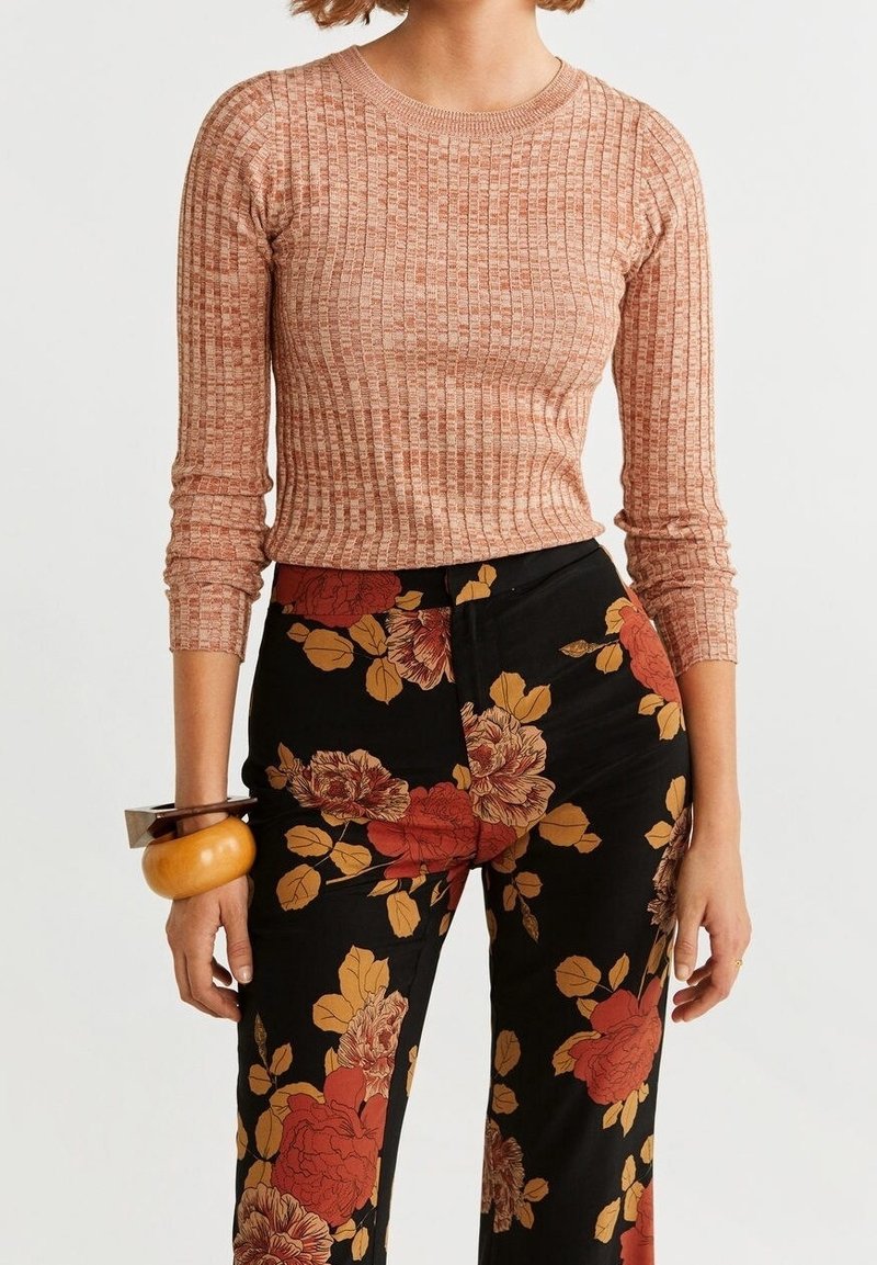 Femme portant un haut à manches longues côtelé, marron clair, et un pantalon noir avec de grands motifs floraux rouges et jaunes, accessoirisée de bracelets épais.