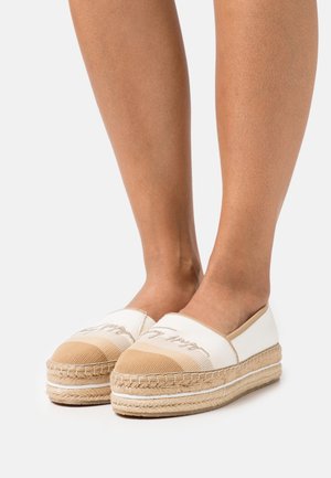 Espadrilles blanches et beiges avec une semelle en jute tissée, présentant un bout arrondi et des détails brodés sur le dessus. Texture de tissu lisse.