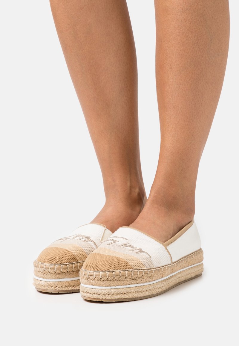 Espadrilles blanches et beiges avec une semelle en jute tissée, présentant un bout arrondi et des détails brodés sur le dessus. Texture de tissu lisse.