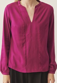 Blusa fucsia con maniche lunghe, scollatura a V e sottili pieghe verticali. Realizzata in tessuto liscio, presenta un'unica tasca sul lato sinistro.
