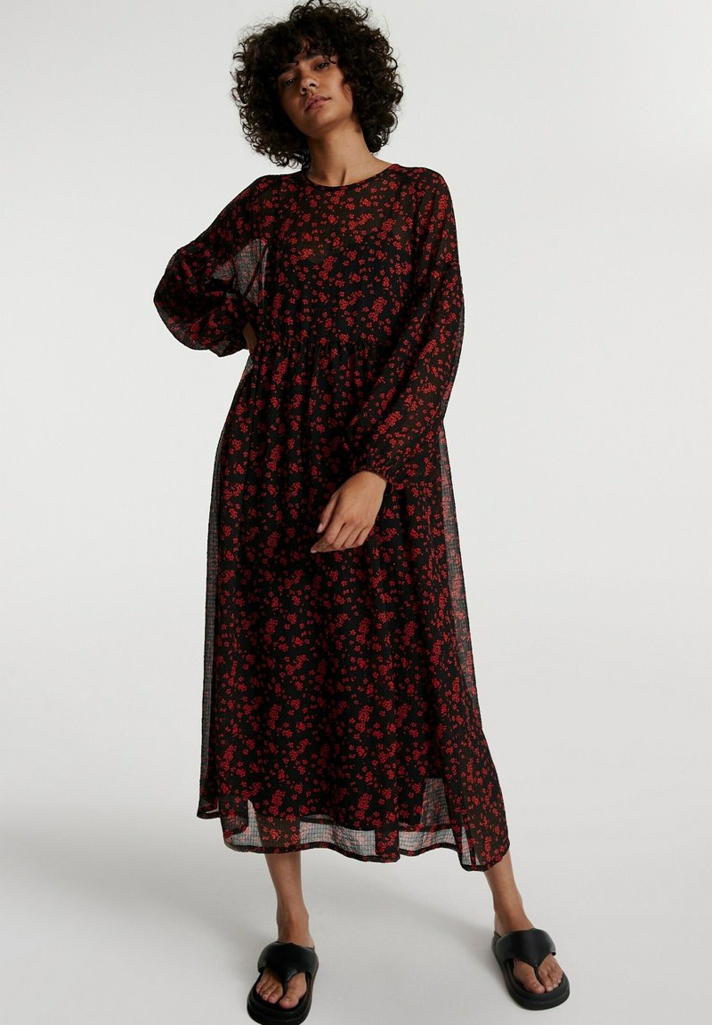 Kleid aus transparenter schwarzer Stoff mit rotem Blumenmuster, lange Ärmel, lockere Passform, schichtetes Design, knielang, getragen mit schwarzen Sandalen.