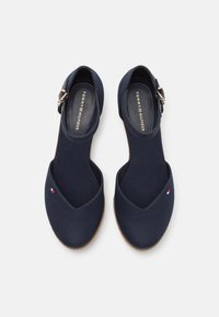 Ett par marinblå Tommy Hilfiger ballerinas för kvinnor med ankelsnören och en liten logotyp på tåområdet, visade uppifrån på en vit bakgrund.