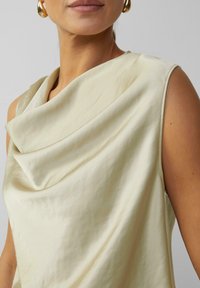 Ärmelloses Oberteil mit Kragen in hellem Beige, das eine weiche Textur, ein drapiertes Design und einen dezenten Glanz aufweist; seitliche Knopfdetails sichtbar.