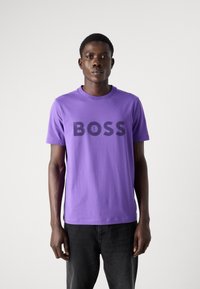 Fialové bavlněné tričko s velkým černým potiskem "BOSS" na přední straně. Krátké rukávy a kulatý výstřih. Nosené s černými džíny.