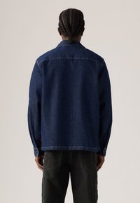 Veste en denim bleu foncé avec col, manches longues et poignets boutonnés, présentant une coupe droite et des détails de couture visibles.