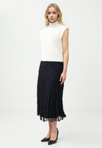 Donna che indossa un maglione senza maniche bianco a collo alto, una gonna midi nera in pizzo e tacchi neri, in piedi davanti a uno sfondo bianco semplice.