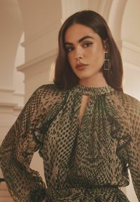 Bluză verde și neagră cu un model cu volane și decolteu tip cheie. Modelul poartă cercei geometrice negri. Textură moale și croi lejer.
