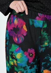 Barevné kalhoty s květinovým potiskem na černém pozadí, s elastickým pasem a hladkou texturou látky s živými akcenty v růžové, zelené a modré barvě.