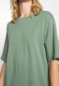 Blondyn ubrany w oversize'owy, szałwiowozielony t-shirt z krótkim rękawem i okrągłym dekoltem na białym tle.