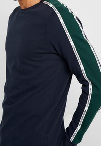 Navyblaues Langarmshirt mit grünen und weißen gestreiften Akzenten an den Ärmeln, mit Logotext und rundem Halsausschnitt.