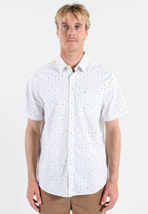 Homme blond portant une chemise blanche à manches courtes avec de petits motifs à points noirs et bleus, et un pantalon marron, debout devant un fond uni.