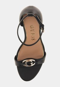 Schwarze Sandale mit offenem Zehenbereich, Knöchelriemen und metallischem "G"-Logo auf dem vorderen Riemen, hellbraune Innensohle mit "GUESS"-Markierung und goldenen Akzentdetails.