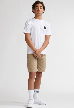 Wit T-shirt met korte mouwen en een zwarte logopatch, gecombineerd met kaki korte broek, witte sneakers en witte sokken met blauwe strepen.