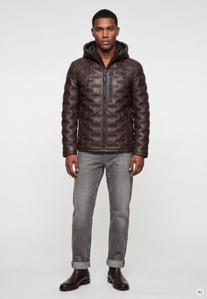 Mann trägt eine braune gesteppte Kapuzenjacke, graue hochgekrempelte Jeans und dunkelbraune Knöchelstiefel und steht vor einem einfarbigen Hintergrund.
