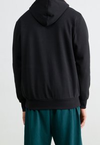 Sudadera negra con capucha ajustable, puños acanalados y cinturilla, vista desde la parte posterior. Combinada con pantalones holgados en color teal.