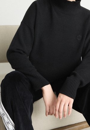 Person sitzt und trägt einen schwarzen, gerippten Rollkragenpullover sowie schwarze Hosen mit weißen Seitenstreifen, auf denen Text zu sehen ist, die Hände ruhen auf den Knien.