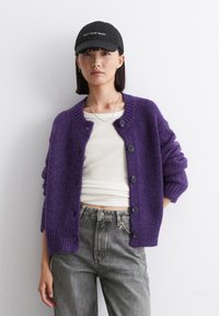Cardigan di lana viola con colletto e polsini a costine, dotato di bottoni neri sul davanti. Indossato su una camicia bianca e jeans grigi.