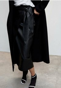 Jupe enveloppante en cuir noir avec un détail de nœud noué à la taille, arborant une texture lisse et soyeuse. Associée à des bottes à talons noires et blanches.