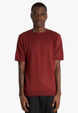 Giovane uomo con capelli corti che indossa una maglietta rossa tinta unita a maniche corte, pantaloni neri, collana a catena e braccialetto, in piedi su uno sfondo bianco.