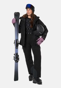 Frau in schwarzer Skibekleidung und lila Handschuhen, die Skier in einer Hand und einen schwarzen Helm in der anderen hält, mit Skibrille auf dem Kopf.