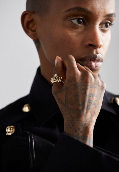 Gold-tone ring with a textured design and clear gemstone, worn on a tattooed hand near a black jacket with gold hardware details.