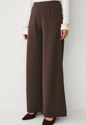 Brune bukser med høj talje og vidde i benene, kombineret med sorte spidse sko og en hvid langærmet top, med guldarmbånd på håndleddene.